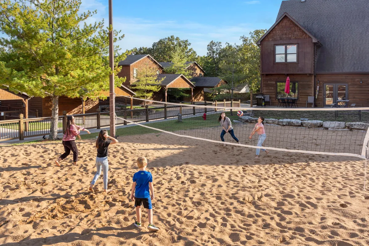 Children play an enthusiastic beach volleyball match on a sand court, surrounded by green spaces and cabins. The scene is lively, with spectators and participants of all ages, encapsulating active outdoor play and resort camaraderie.