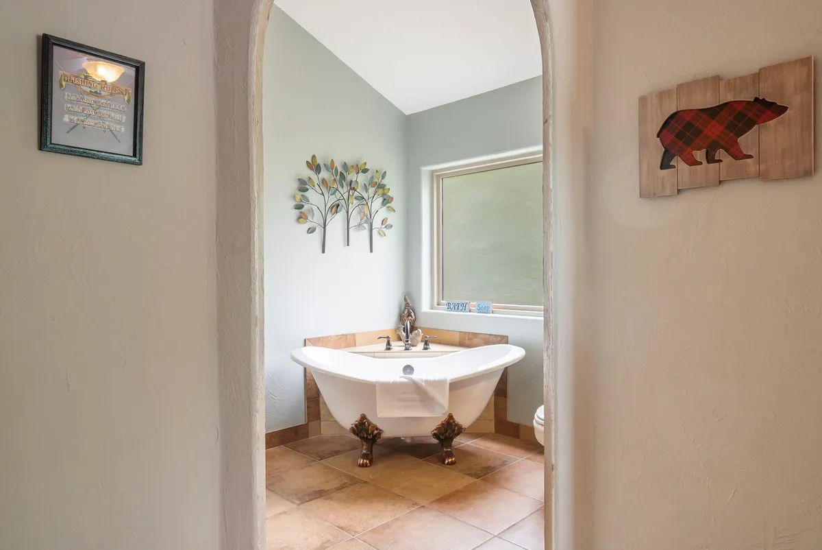 A serene ensuite bathroom featuring a freestanding clawfoot-style bathtub, decorative tree and bear-themed wall art, and tranquil lighting. The space exudes a spa-like feel perfect for unwinding after a day of lakeside adventures.