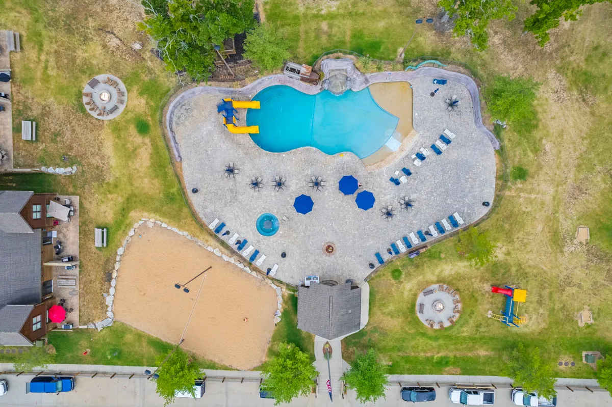 This vibrant exterior scene shows a sparkling swimming pool with a water slide, a sand volleyball court, several seating areas shaded by umbrellas, and a fire pit nearby. Families and children are enjoying the various amenities, creating a lively, resort-like atmosphere.