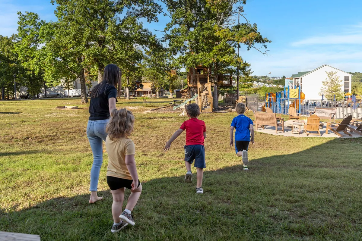 Children are seen running and playing on a colorful playground and treehouse surrounded by nature, with a seating area nearby for families. The mood is joyful and carefree, emphasizing outdoor play and family bonding.