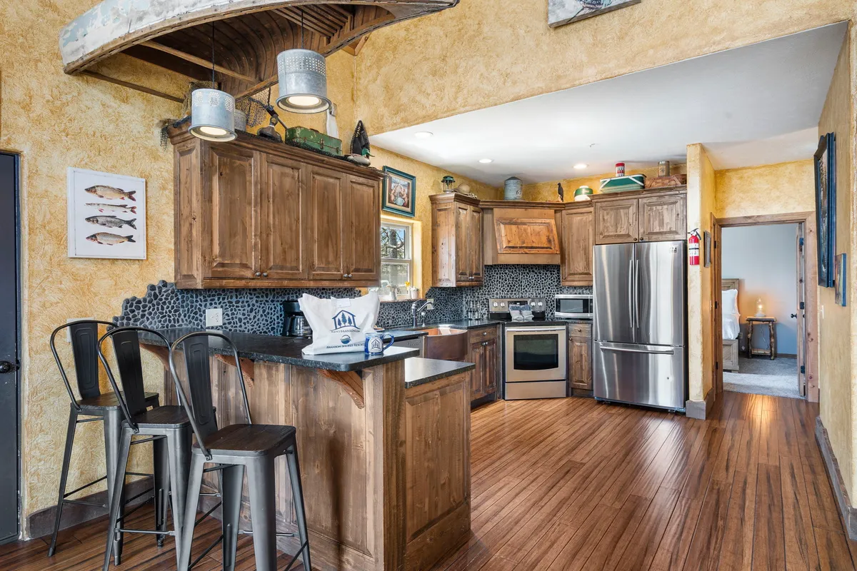 A detail-rich kitchen view featuring stainless steel appliances, wooden cabinets, bar stools along a counter, and decorative artwork. The stylish stone backsplash is visible, and the space exudes warmth and hospitality ideal for shared meal prep and gatherings.