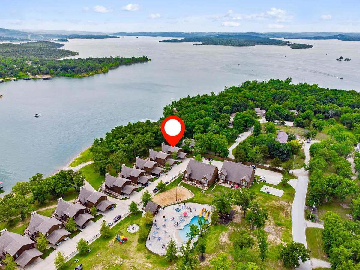 A picturesque lakeside scene including several cabins, a playground, a swimming pool, and the tranquil lake itself. The setting exudes a relaxed, recreational atmosphere, with amenities for both active play and calm relaxation. The communal spaces invite group activities from swimming to shared meals on the porch, all set against a beautiful lakeside backdrop.