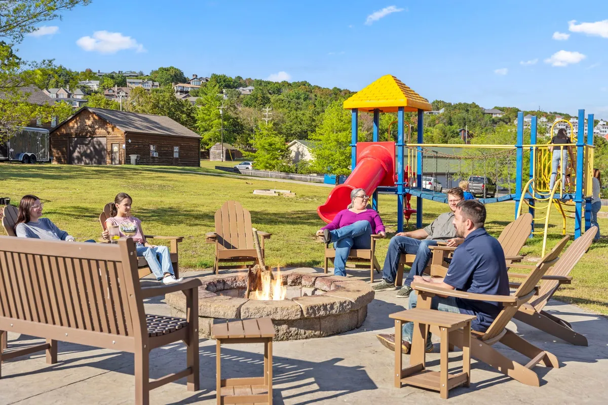 An exterior gathering area featuring a fire pit surrounded by seating, with playground equipment nearby and people gathered around the fire. The open grassy area adds a communal and playful atmosphere, great for socializing and relaxing with friends, family, and pets under the stars.