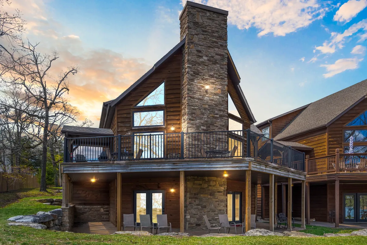 The hero image showcases the inviting exterior of a spacious lodge-style home nestled on the edge of Table Rock Lake. A large stone chimney hints at a cozy indoor fireplace, while generous balconies with comfortable seating invite guests to unwind and soak in breathtaking sunset views. The scene exudes warmth, with the soft glow of dusk reflecting on the water and spacious decks promising memorable outdoor meals and relaxation.
