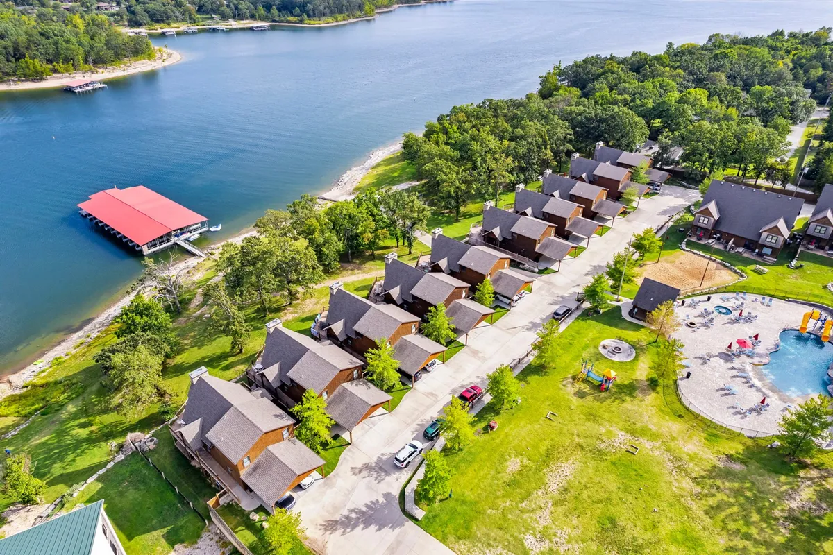 A broad exterior view of the resort, featuring a row of cabins, the swimming pool, a play area, and a dock with a red roof. The shot captures multiple communal amenities and the natural landscape, suggesting both relaxation and recreation.