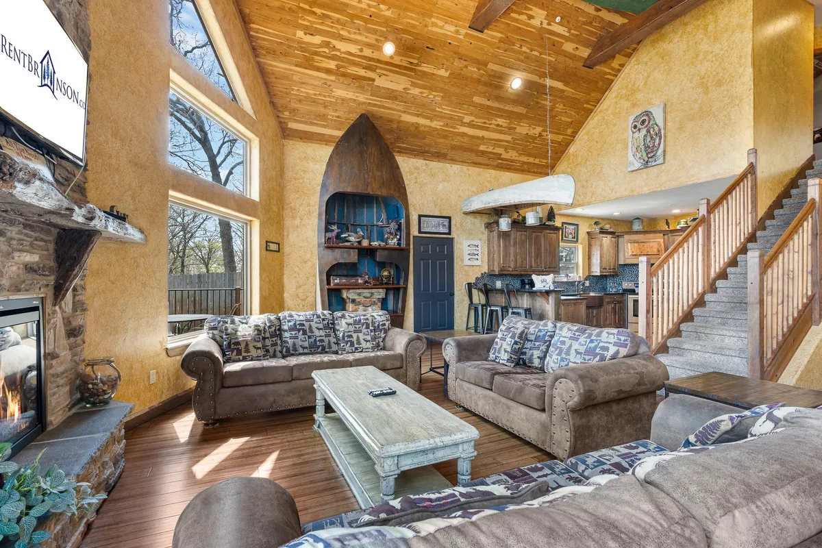 A cozy living and dining space with a large stone fireplace, expansive windows overlooking the lake, wooden beams overhead, and unique boat decor suspended from the ceiling. The arrangement encourages both group and intimate gatherings, with lakeside charm throughout.