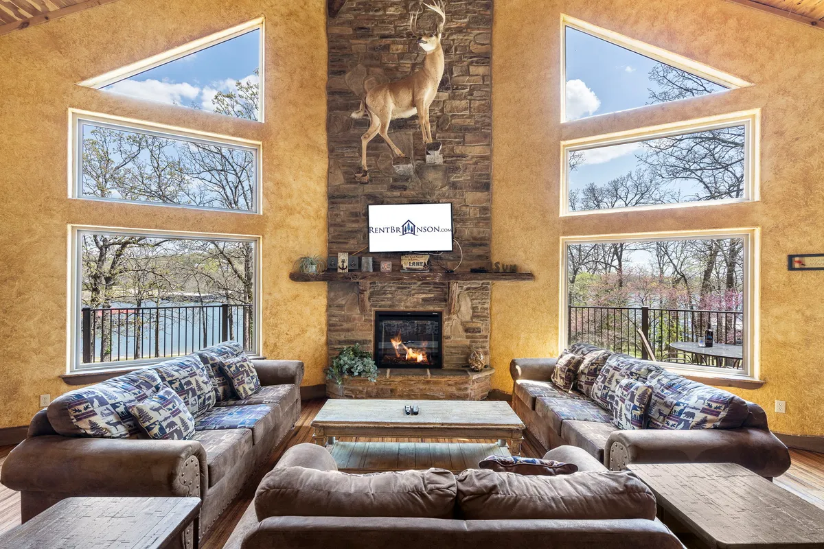 This image features the great room with a large stone fireplace, comfortable sofas, spacious windows overlooking Table Rock Lake, and a warm, welcoming atmosphere. The vaulted ceiling and expansive glass provide an immediate visual connection to the lake just outside.