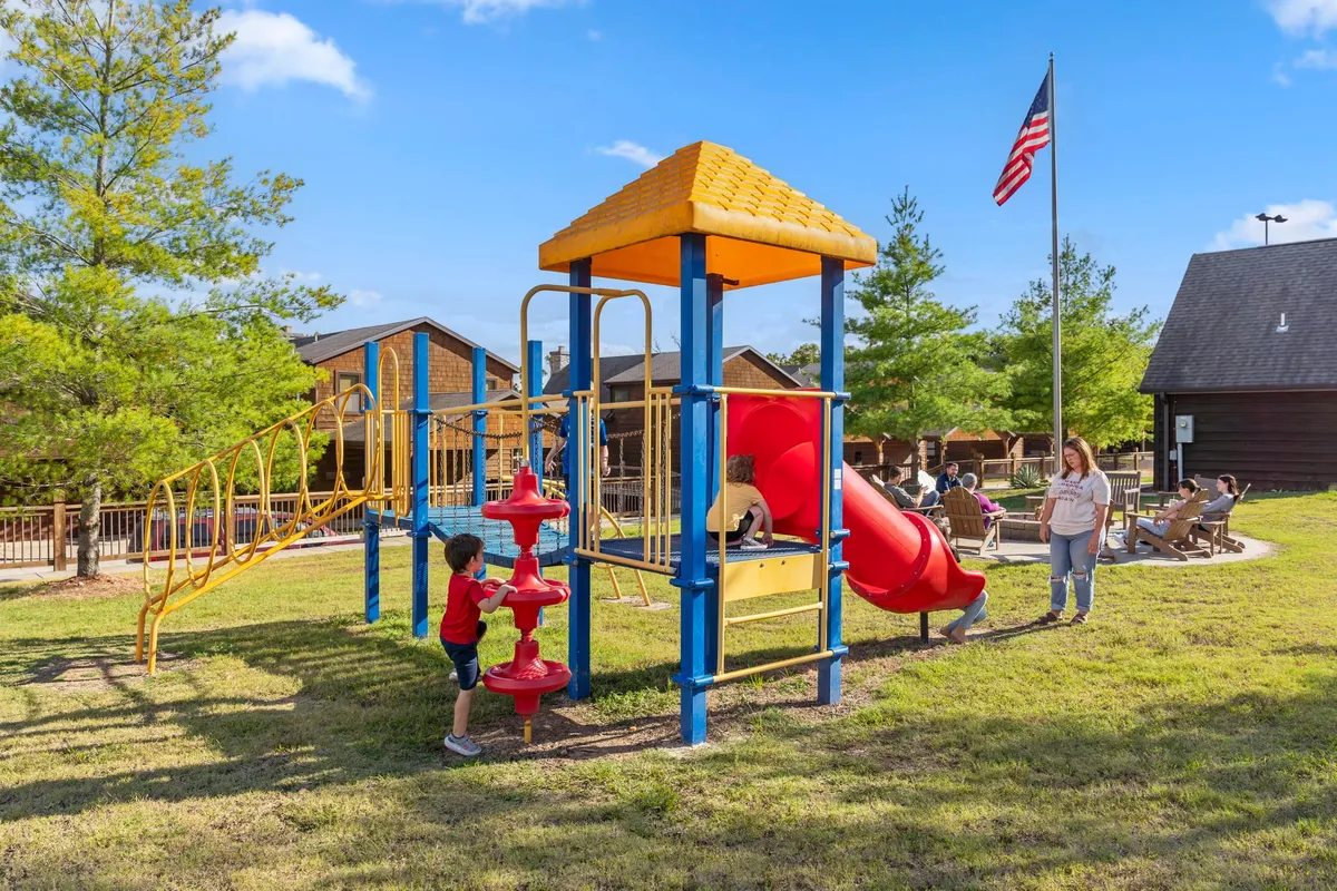 A lively playground area where children are playing on equipment while adults relax nearby. The setting is open, sunny, and framed by greenery, capturing the joy and energy of families enjoying outdoor activities together.