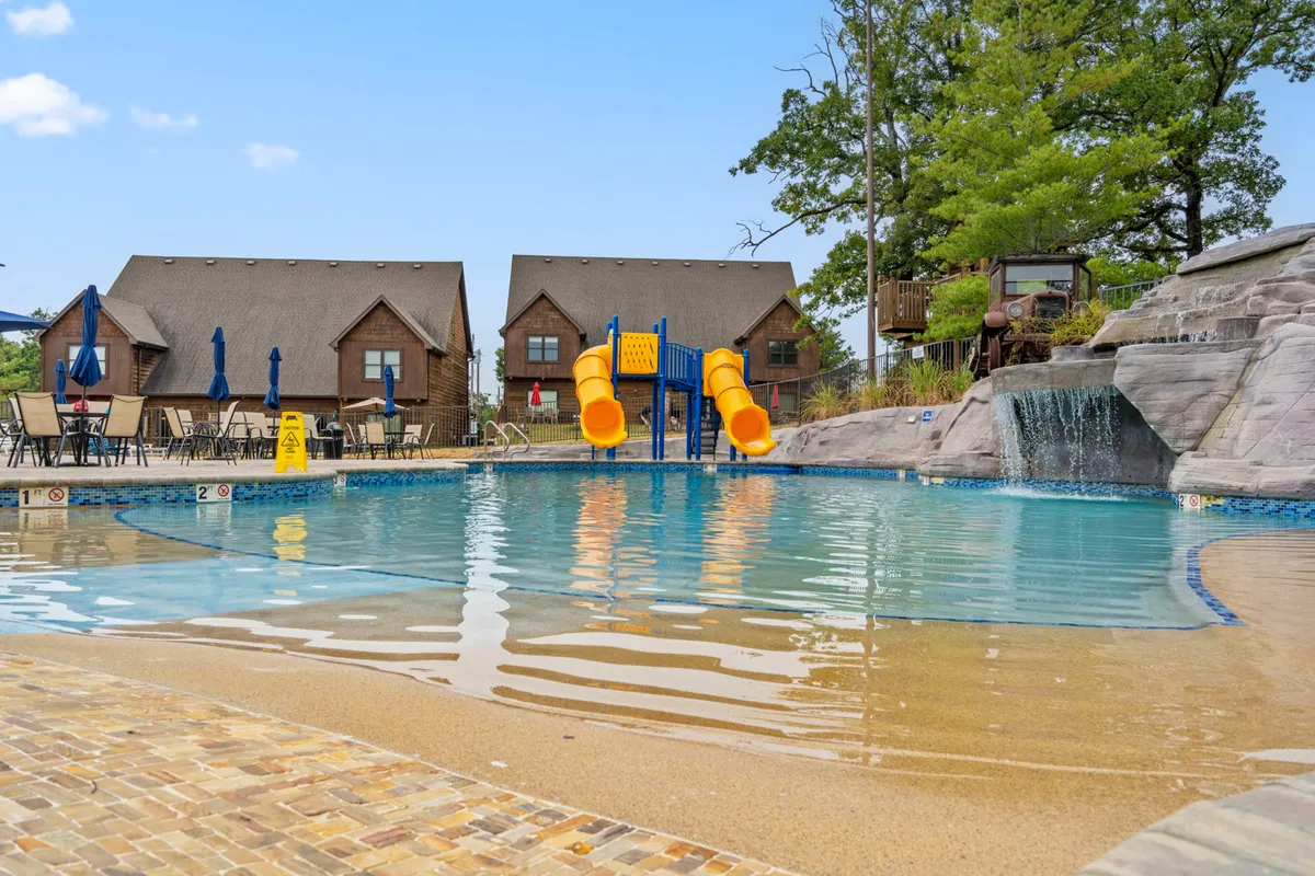 A vibrant outdoor pool scene featuring a swimming pool with a waterslide, lounge chairs, and a cascading waterfall. Children can be seen playing in the water while families relax poolside, enjoying the sunny weather and cheerful atmosphere.