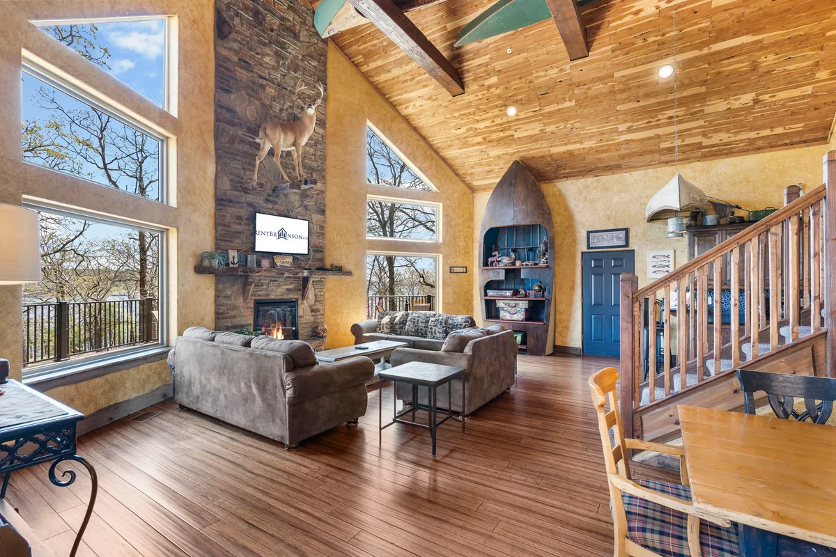 A sweeping view of the living and dining areas highlights the rustic dining table, large stone fireplace, wooden beams, expansive windows, and decorative touches like a deer mount. The layout flows gracefully towards glass doors and outdoor views, reinforcing the sense of space and connection.